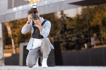 Young happy male photographer with professional camera taking photo while sitting on city street