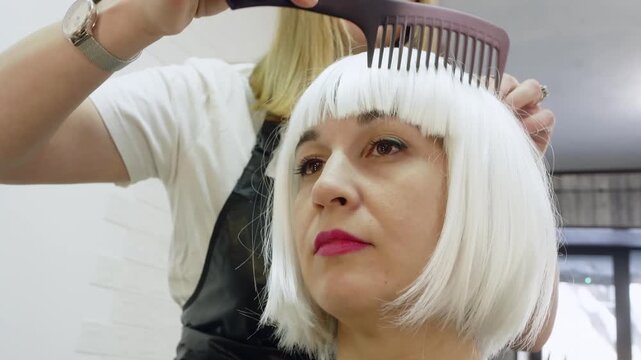 Focused senior woman with short white bob haircut receiving styling with large comb from female hairdresser in bright salon, symbol of care, fashion and beauty.