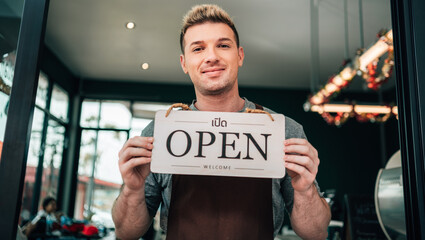 A smiling man in an apron stands in the doorway of his cafe, holding up a wooden "OPEN" sign. The sign also has Thai text and the word "WELCOME."