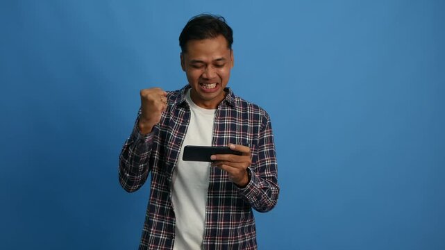 Happy asian man having fun playing online mobile game on smartphone while standing over blue background
