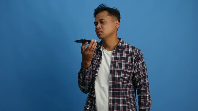 Young Asian man standing while on phone with loud speaker mode. Isolated on blue background