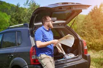Man tourist leans on open trunk of SUV, deeply focused on map. Traveler wears blue shirt and beige pants, standing in lush, green area, carefully planning or navigating his route.