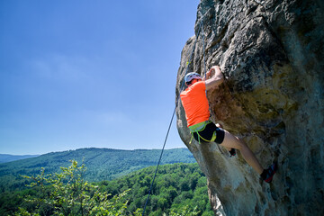 Aerial view of male rock climber ascending rugged limestone cliff with harness and rope for safety. Sportsman climbing on vertical large boulder at Dobvush Rocks in Carpathian mountains, Ukraine.