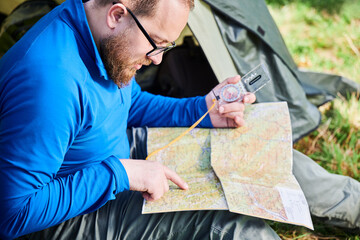 Close up man in glasses closely examines map while holding compass. He sits by his tent, deeply focused on navigation. Lush greenery, suggesting serene outdoor adventure.