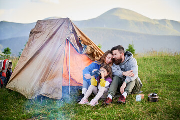 Family of three sits together in front of orange tent in grassy mountain campsite. Child uses binoculars while parents share tender moment. Peaceful and intimate outdoor experience.