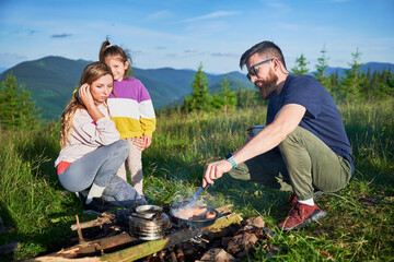 Man tourist cooking over campfire while woman and young girl standing nearby in picturesque mountain meadow. Father tending to food. Lush greenery and rolling mountains under clear blue sky.