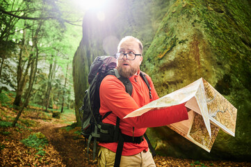 Bearded tourist man looks puzzled, studies map while stands in front of large rock in dense forest. Traveler with grey backpack and glasses, trying to figure out his direction.