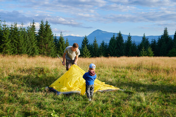 Man hiker and young boy work together to set up tent on grassy hillside. Pine trees and distant mountains under partly cloudy sky.