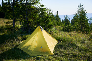 Orange tourist tent on grassy hillside under sunset sky. Surrounded by young pine trees. Stunning view of distant mountains and creates peaceful and picturesque camping spot in the morning.
