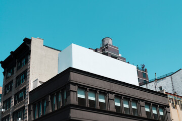 A large blank billboard stands on top of city buildings under a clear blue sky, surrounded by urban architecture and windows, ready for advertisements or creative displays.