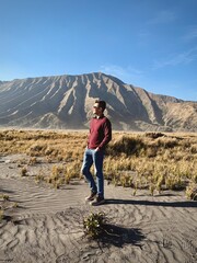 A guy posing in the middle of sea of sand and savannah with hills and mountain at the back. Located in the Mount Bromo caldera, at Bromo Tengger Semeru National park. Beauty of nature.