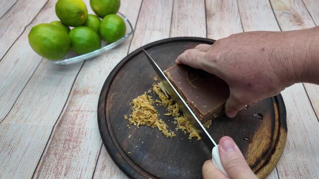 close-up hand using a kitchen knife to cut and shave hard panela (raw sugar cane block) on a dark wooden board, with fresh green limes visible in the background for a traditional beverage preparation.