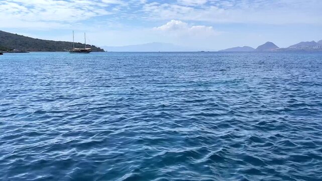 A stunning view of deep blue sea with gentle waves, a traditional gulet near a rocky coastline, and hazy mountains in the distance under a partly cloudy sky.