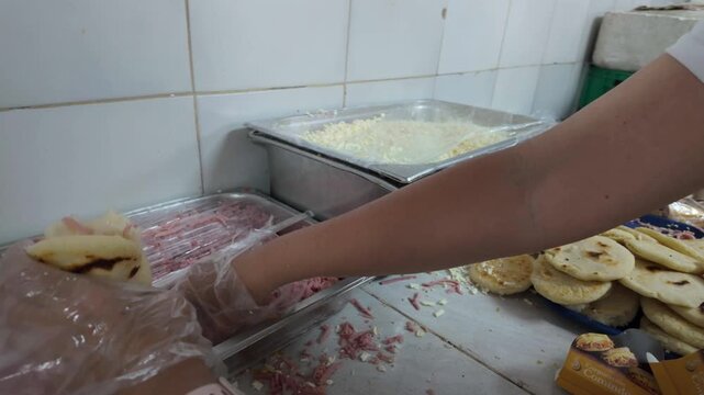close-up food service worker's gloved hands preparing Venezuelan arepas in a commercial setting. Hands are seen scooping shredded ham (jam&oacute;n) and cheese from metal containers to stuff arepas