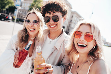 Man and two young beautiful smiling hipster female. Carefree women posing outdoors. Positive models holding and drinking fresh cocktail drink in glass bottle. In the street. Cheerful and happy