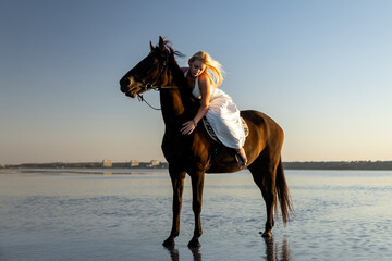 A woman in a flowing white dress leans affectionately on her horse while standing in the calm waters of Kuyalnik Lake at sunset.