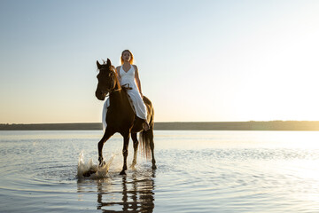 A woman in a white dress rides her horse through the shallow waters of Kuyalnik Lake, illuminated by the warm sunset glow.