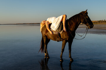 A woman in a white dress reclines gracefully across her horse, bathed in golden sunset light at Kuyalnik Lake.