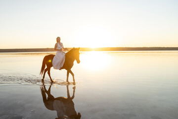 A woman in a white dress rides her horse across Kuyalnik Lake at sunset, glowing light reflecting off the still water.