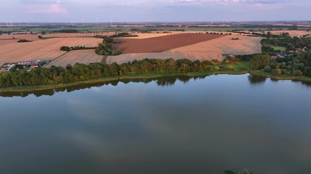 Aerial view of a placid lake reflecting the sky, contrasting with the adjacent harvested fields and distant wind turbines, Battin, Brandenburg, Germany.