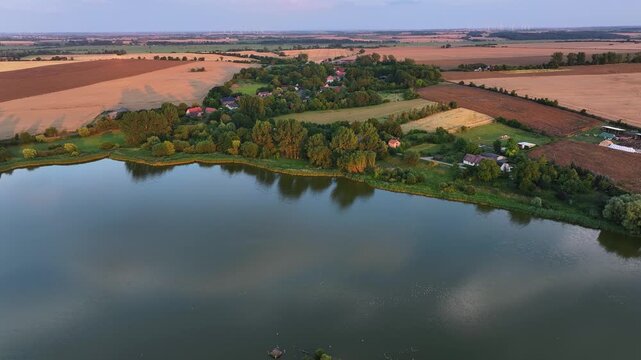 Aerial view of the tranquil lake reflecting the sky, bordered by a village and fields displaying a mosaic of golden and green hues, Battin, Brandenburg, Germany.