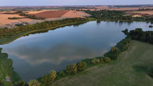 Aerial view of a tranquil lake reflecting the sky, bordered by lush green trees and golden fields, creating a serene contrast, Battin, Brandenburg, Germany.
