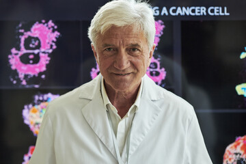 Portrait of confident senior Caucasian male doctor smiling and standing in front of scientific display showing cancer cell research images, wearing lab coat, presenting medical data at conference
