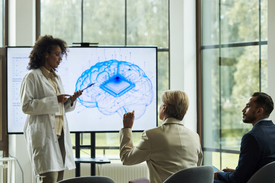 Young Black female doctor explaining brain microchip concept for neurological diseases treatment to Caucasian colleagues in conference room with digital diagram displayed on screen