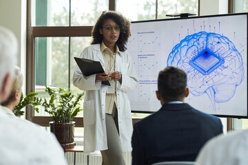 Young Black woman presenting neuroscience research to diverse group, standing in front of digital brain diagram with microchip illustration, holding clipboard, wearing lab coat