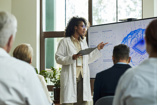 Young Black woman presenting scientific data to group of middle aged and senior Caucasian men in conference room, standing near digital screen displaying brain diagram and charts - Powered by Adobe