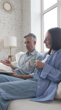 Middle-aged man and woman in casual clothes sitting on sofa with laptop, drinking coffee and discussing at home. Concept of teamwork, communication, planning, lifestyle and collaboration