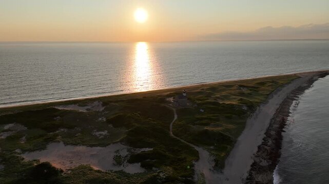 Aerial view of the North Light lighthouse on Block Island, bathed in the warm glow of the setting sun, casting long shadows across the sandy dunes, New Shoreham, Rhode Island, United States.