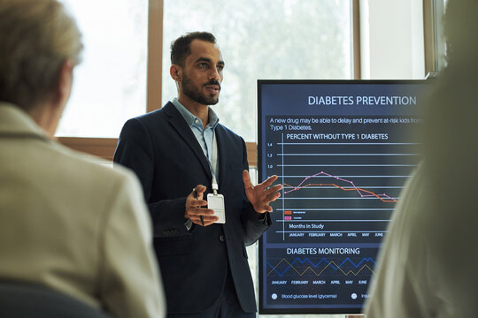 Young biracial man presenting diabetes prevention data to group, gesturing while standing near large digital screen displaying medical statistics and research information - Powered by Adobe
