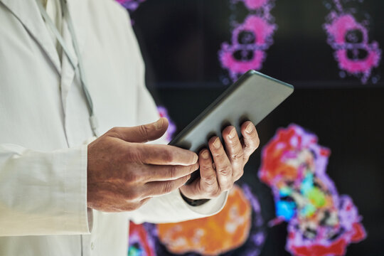 Middle aged Caucasian man in white lab coat holding digital tablet, standing in front of medical imaging display with colorful anatomical scans, using technology for healthcare analysis at conference