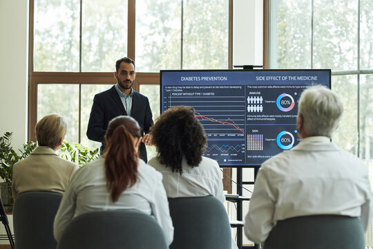 Young Middle Eastern man in suit presenting medical data, on diabetes prevention and medicine side effects, to group of senior and middle aged multiethnic doctors seated in conference room