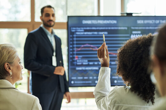 Young male researcher presenting diabetes prevention data on large screen, while Black young woman raising hand to ask question, during medical seminar with diverse audience