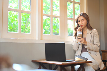 Young woman in beige suit sitting by window in cafe, drinking iced coffee and working on laptop
