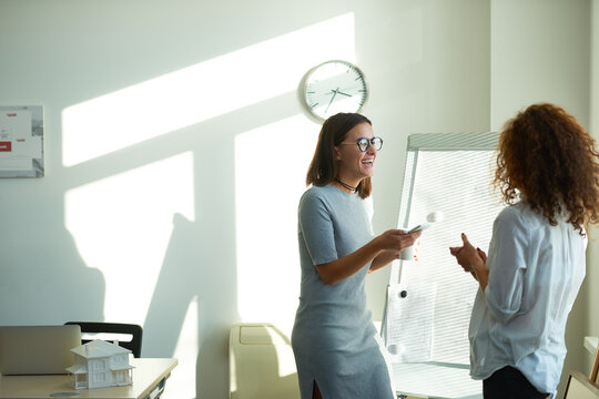 Young stylish woman in gray dress and round eyeglasses with smartphone in hands, laughing cheerfully after getting funny message and sharing it with her curly colleague - Powered by Adobe