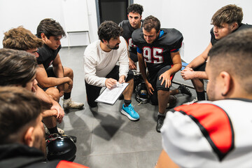American football coach discussing game strategy and tactics with players in a locker room, emphasizing teamwork and leadership