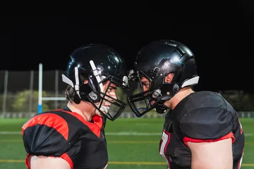 Fototapeten Fußball American football players in helmets and shoulder pads facing each other intensely on a stadium field at night, symbolizing rivalry and determination  © Koldo_Studio