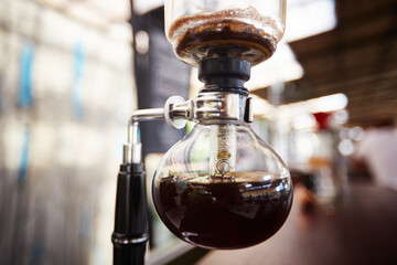 Close up showing glass siphon coffee maker brewing fresh coffee in modern cafe setting, focusing on lower chamber with dark liquid and visible brewing process, no people visible