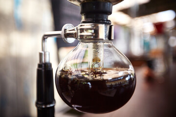 Close up showing glass siphon coffee maker brewing fresh coffee, transparent round vessel with dark liquid and visible condensation, laboratory style equipment in use