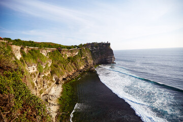 Rocky coastal cliff stretching along shoreline with ocean waves crashing against base, green vegetation covering top of cliff, blue sky meeting horizon, no people visible