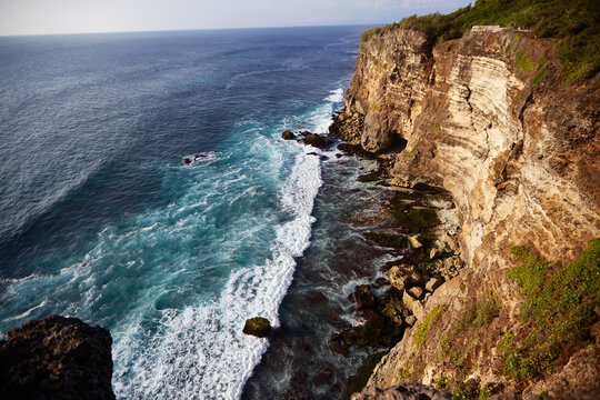 Rocky coastline with steep cliffs meeting ocean waves, rugged shoreline stretching into distance, blue water crashing against rocks, natural landscape with dramatic elevation