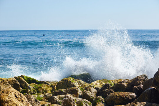 Ocean waves crashing against rocky shoreline with water splashing high, clear blue sky in background, natural seascape showing dynamic movement and coastal landscape