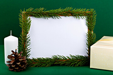 Mockup. Christmas. Festive mockup featuring blank frame for design or advertising surrounded by pine branches, white candle, pine cone and gift box against rich green background, ideal for holiday
