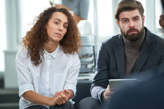 Stylish curly woman in white shirt and bearded man in suit and turtleneck sitting in modern office, listening to their partners with serious face expressions