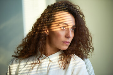 Close-up portrait of brown-eyed woman in pure white buttoned up shirt sitting in office and looking to the side with sentimental expression. Light lines from Venetians blinds seen on her forehead