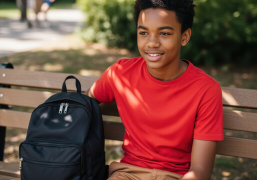 Smiling teenage boy in a red shirt sitting on a park bench with a black backpack during a sunny day outdoors - Powered by Adobe