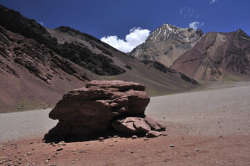 Orange red sand desert, rocky formations and mountains background, blue sky above. A large rock in the middle of the desert